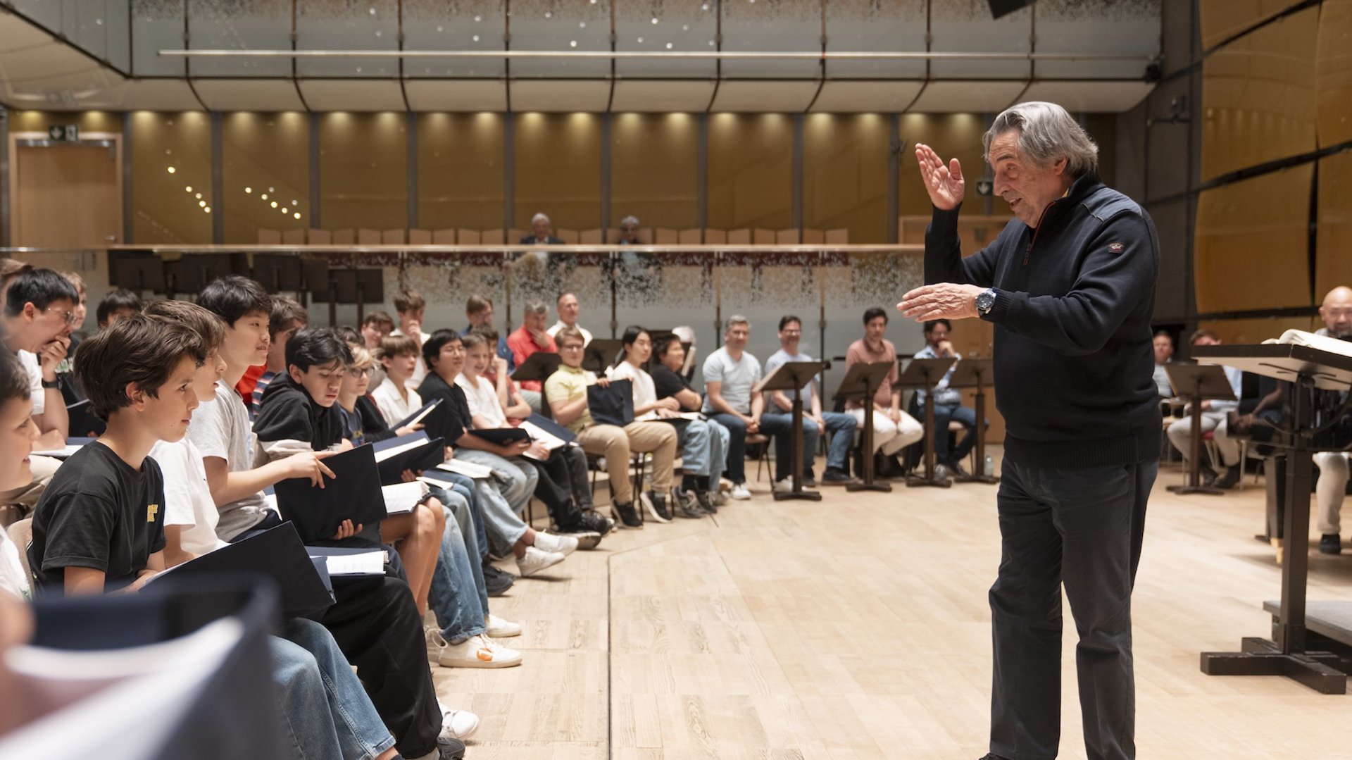 Eine Fotografie von Lukas Beck im Gläsernen Saal (Magna Auditorium) des Wiener Musikvereins. Das Bild zeigt eine hochempathische Probensituation: Der Stardirigent Riccardo Muti steht im dunklen Pullover im Profil rechts auf dem Holzparkett und dirigiert mit schwungvoll erhobenen Händen. Die Sängerknaben sitzen links von ihm im Halbkreis aufgereiht, halten ihre Notenmappen auf den Knien und blicken hochkonzentriert zum Maestro. Die moderne, akustisch optimierte Holz- und Glasarchitektur des Saals ist im Hintergrund deutlich sichtbar. Gemäß DSGVO werden keine Namen der Kinder genannt.