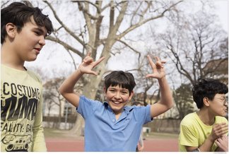 Eine Fotografie von Lukas Beck auf dem Allwetter-Sportplatz des Campus Augarten. Zu sehen ist eine fröhliche Freizeitszene: Ein junger Sängerknabe im blauen Poloshirt steht im Zentrum, blickt lachend direkt in die Kamera und formt mit beiden Händen scherzhafte Gesten über seinem Kopf. Links von ihm steht ein Mitschüler im hellen T-Shirt im Profil, rechts ist ein weiterer Junge im gelben Oberteil angeschnitten. Im Hintergrund erstrecken sich die großen Bäume des Parks unter leicht bewölktem Himmel. Gemäß DSGVO werden keine Namen der Kinder genannt.