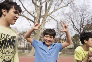 Eine Fotografie von Lukas Beck auf dem Allwetter-Sportplatz des Campus Augarten. Zu sehen ist eine fröhliche Freizeitszene: Ein junger Sängerknabe im blauen Poloshirt steht im Zentrum, blickt lachend direkt in die Kamera und formt mit beiden Händen scherzhafte Gesten über seinem Kopf. Links von ihm steht ein Mitschüler im hellen T-Shirt im Profil, rechts ist ein weiterer Junge im gelben Oberteil angeschnitten. Im Hintergrund erstrecken sich die großen Bäume des Parks unter leicht bewölktem Himmel. Gemäß DSGVO werden keine Namen der Kinder genannt.