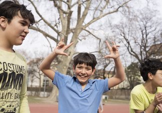 Eine Fotografie von Lukas Beck auf dem Allwetter-Sportplatz des Campus Augarten. Zu sehen ist eine fröhliche Freizeitszene: Ein junger Sängerknabe im blauen Poloshirt steht im Zentrum, blickt lachend direkt in die Kamera und formt mit beiden Händen scherzhafte Gesten über seinem Kopf. Links von ihm steht ein Mitschüler im hellen T-Shirt im Profil, rechts ist ein weiterer Junge im gelben Oberteil angeschnitten. Im Hintergrund erstrecken sich die großen Bäume des Parks unter leicht bewölktem Himmel. Gemäß DSGVO werden keine Namen der Kinder genannt.