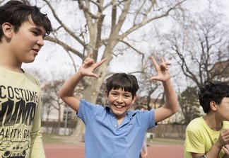 Eine Fotografie von Lukas Beck auf dem Allwetter-Sportplatz des Campus Augarten. Zu sehen ist eine fröhliche Freizeitszene: Ein junger Sängerknabe im blauen Poloshirt steht im Zentrum, blickt lachend direkt in die Kamera und formt mit beiden Händen scherzhafte Gesten über seinem Kopf. Links von ihm steht ein Mitschüler im hellen T-Shirt im Profil, rechts ist ein weiterer Junge im gelben Oberteil angeschnitten. Im Hintergrund erstrecken sich die großen Bäume des Parks unter leicht bewölktem Himmel. Gemäß DSGVO werden keine Namen der Kinder genannt.