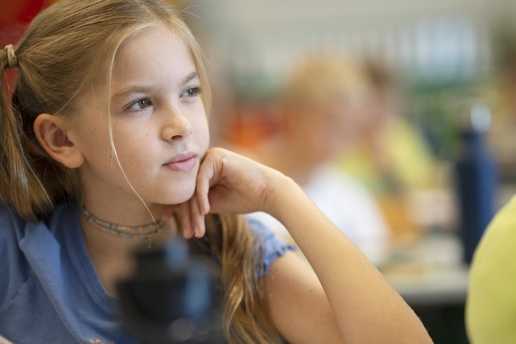 Ein ausdrucksstarkes Porträt einer Schülerin aus der 1. Klasse des Gymnasiums der Wiener Sängerknaben. Das Mädchen gehört den Wiener Chormädchen an. Sie hat lange, blonde Haare, die teilweise seitlich zurückgebunden sind, trägt ein hellblaues T-Shirt und hat ihr Kinn nachdenklich auf ihre linke Hand gestützt. Ihr Blick ist aufmerksam zur Seite gerichtet, während sie konzentriert dem Unterricht folgt. Die Aufnahme von Lukas Beck arbeitet mit einer sehr geringen Schärfentiefe: Im unscharfen Vordergrund ist eine dunkle Trinkflasche zu erkennen, während der Hintergrund mit Mitschülern im Klassenzimmer sanft verschwimmt. Das natürliche Licht setzt das Gesicht der jungen Sängerin klar in Szene. Urheberrecht: © Lukas Beck.
