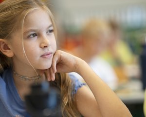 Ein ausdrucksstarkes Porträt einer Schülerin aus der 1. Klasse des Gymnasiums der Wiener Sängerknaben. Das Mädchen gehört den Wiener Chormädchen an. Sie hat lange, blonde Haare, die teilweise seitlich zurückgebunden sind, trägt ein hellblaues T-Shirt und hat ihr Kinn nachdenklich auf ihre linke Hand gestützt. Ihr Blick ist aufmerksam zur Seite gerichtet, während sie konzentriert dem Unterricht folgt. Die Aufnahme von Lukas Beck arbeitet mit einer sehr geringen Schärfentiefe: Im unscharfen Vordergrund ist eine dunkle Trinkflasche zu erkennen, während der Hintergrund mit Mitschülern im Klassenzimmer sanft verschwimmt. Das natürliche Licht setzt das Gesicht der jungen Sängerin klar in Szene. Urheberrecht: © Lukas Beck.