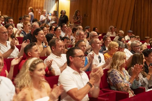 Eine Fotografie von Lukas Beck, die einen Teil des Publikums im MuTh (Konzertsaal der Wiener Sängerknaben) zeigt. Anlass ist die feierliche Uniformübergabe. Im Bild sind Eltern und Geschwister zu sehen, die in den roten Polstersesseln des Saals sitzen, fröhlich blicken und enthusiastisch Beifall klatschen. Die Wände des Saals sind mit vertikalen Holzpaneelen verkleidet.