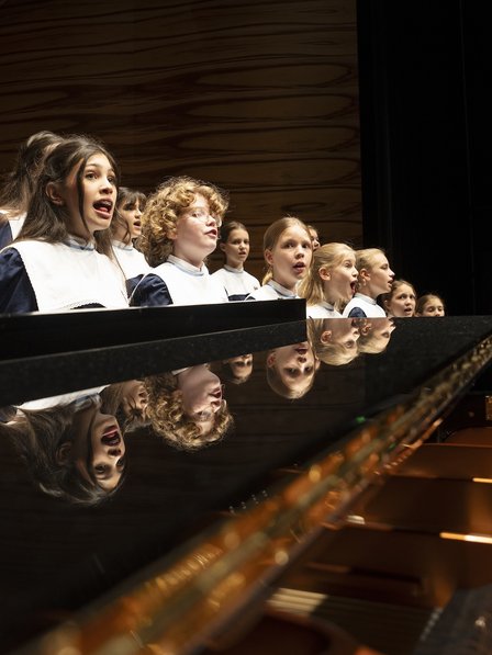 Ein Mädchen-Chor in Uniform singt neben einem Klavier.
