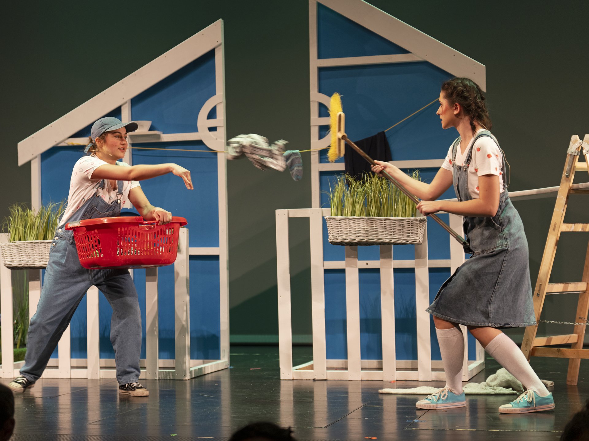 Eine Fotografie von Lukas Beck im MuTh anlässlich der Aufführung von Engelbert Humperdincks Oper „Hänsel und Gretel“ im Jahr 2025. Das Bild zeigt eine dynamische Probenszene mit zwei Sängerinnen des Chorus Juventus in den Titelrollen: Links steht Hänsel in Jeanslatzhose und Cap, hält eine rote Wäschewanne und bewirft Gretel schwungvoll mit einem weißen Kleidungsstück. Rechts wehrt sich Gretel (ebenfalls in Latzhose und mit Zöpfen) scherzhaft mit einem Besen. Im Hintergrund dient eine stilisierte, blau-weiße Konstruktion als Kulisse für das Haus der Eltern. Rechts ist eine Holzleiter zu sehen. Aus DSGVO-Gründen werden die Namen der Sängerinnen nicht genannt.