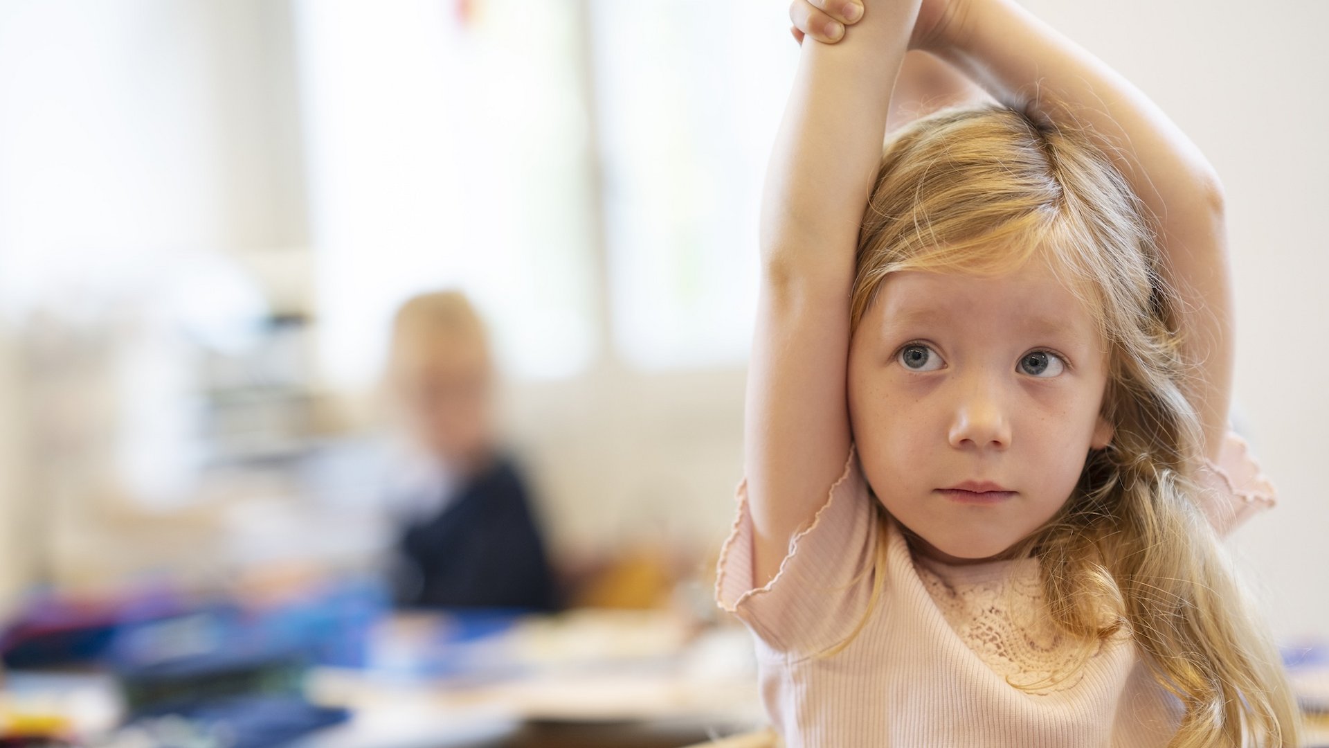 Ein junges Mädchen sitzt im Klassenraum und zeigt mit beiden Armen auf.