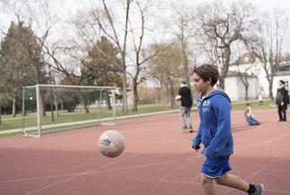 Eine Fotografie von Lukas Beck auf dem Sportplatz des Campus Augarten. Das Bild zeigt eine lebendige Freizeitszene beim Fußballspielen: Im Vordergrund läuft ein dunkelblonder Junge in einer blauen Sportjacke und kurzen blauen Hosen nach links einem in der Luft fliegenden Fußball hinterher. Links im Hintergrund steht ein Fußballtor aus Metall auf dem roten Allwetterplatz. Im weiteren Hintergrund sind Bäume und zwei Mitschüler zu sehen, von denen einer einen Ball hält. Gemäß DSGVO werden keine Namen der Kinder genannt.