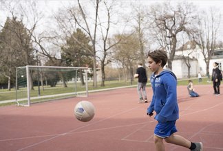 Eine Fotografie von Lukas Beck auf dem Sportplatz des Campus Augarten. Das Bild zeigt eine lebendige Freizeitszene beim Fußballspielen: Im Vordergrund läuft ein dunkelblonder Junge in einer blauen Sportjacke und kurzen blauen Hosen nach links einem in der Luft fliegenden Fußball hinterher. Links im Hintergrund steht ein Fußballtor aus Metall auf dem roten Allwetterplatz. Im weiteren Hintergrund sind Bäume und zwei Mitschüler zu sehen, von denen einer einen Ball hält. Gemäß DSGVO werden keine Namen der Kinder genannt.