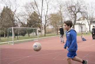 Eine Fotografie von Lukas Beck auf dem Sportplatz des Campus Augarten. Das Bild zeigt eine lebendige Freizeitszene beim Fußballspielen: Im Vordergrund läuft ein dunkelblonder Junge in einer blauen Sportjacke und kurzen blauen Hosen nach links einem in der Luft fliegenden Fußball hinterher. Links im Hintergrund steht ein Fußballtor aus Metall auf dem roten Allwetterplatz. Im weiteren Hintergrund sind Bäume und zwei Mitschüler zu sehen, von denen einer einen Ball hält. Gemäß DSGVO werden keine Namen der Kinder genannt.