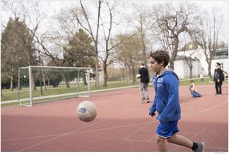 Eine Fotografie von Lukas Beck auf dem Sportplatz des Campus Augarten. Das Bild zeigt eine lebendige Freizeitszene beim Fußballspielen: Im Vordergrund läuft ein dunkelblonder Junge in einer blauen Sportjacke und kurzen blauen Hosen nach links einem in der Luft fliegenden Fußball hinterher. Links im Hintergrund steht ein Fußballtor aus Metall auf dem roten Allwetterplatz. Im weiteren Hintergrund sind Bäume und zwei Mitschüler zu sehen, von denen einer einen Ball hält. Gemäß DSGVO werden keine Namen der Kinder genannt.