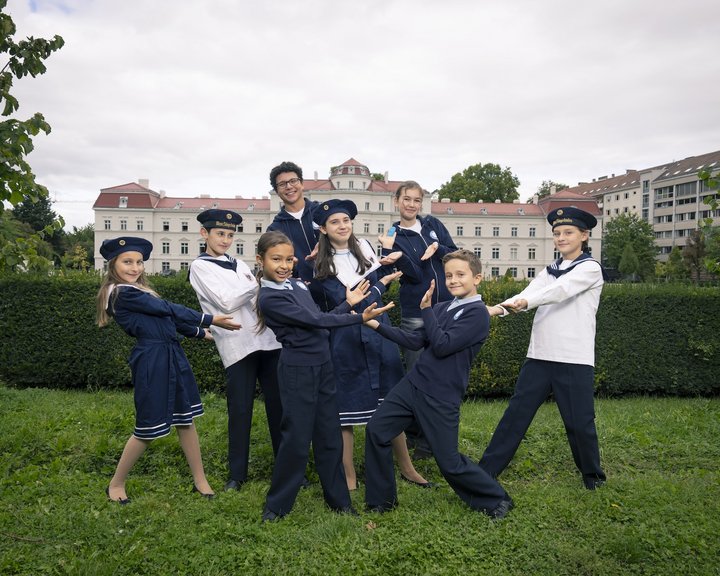 Eine Fotografie von Lukas Beck im Park des Palais Augarten. Das Bild symbolisiert die Vielfalt des Campus: Eine Gruppe von acht jungen Chormitgliedern (Mädchen und Jungen) posiert in einer spielerisch-dynamischen Formation auf einer grünen Rasenfläche. Sie tragen die verschiedenen offiziellen Outfits der Chöre – darunter dunkelblaue Matrosenkleider für die Mädchen sowie die klassischen blauen und weißen Matrosenuniformen der Jungen. Im Hintergrund erstreckt sich eine grüne Hecke, hinter der das barocke Palais Augarten unter leicht bewölktem Himmel zu sehen ist. Gemäß DSGVO werden keine Namen der Kinder genannt.