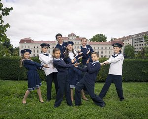 Eine Fotografie von Lukas Beck im Park des Palais Augarten. Das Bild symbolisiert die Vielfalt des Campus: Eine Gruppe von acht jungen Chormitgliedern (Mädchen und Jungen) posiert in einer spielerisch-dynamischen Formation auf einer grünen Rasenfläche. Sie tragen die verschiedenen offiziellen Outfits der Chöre – darunter dunkelblaue Matrosenkleider für die Mädchen sowie die klassischen blauen und weißen Matrosenuniformen der Jungen. Im Hintergrund erstreckt sich eine grüne Hecke, hinter der das barocke Palais Augarten unter leicht bewölktem Himmel zu sehen ist. Gemäß DSGVO werden keine Namen der Kinder genannt.