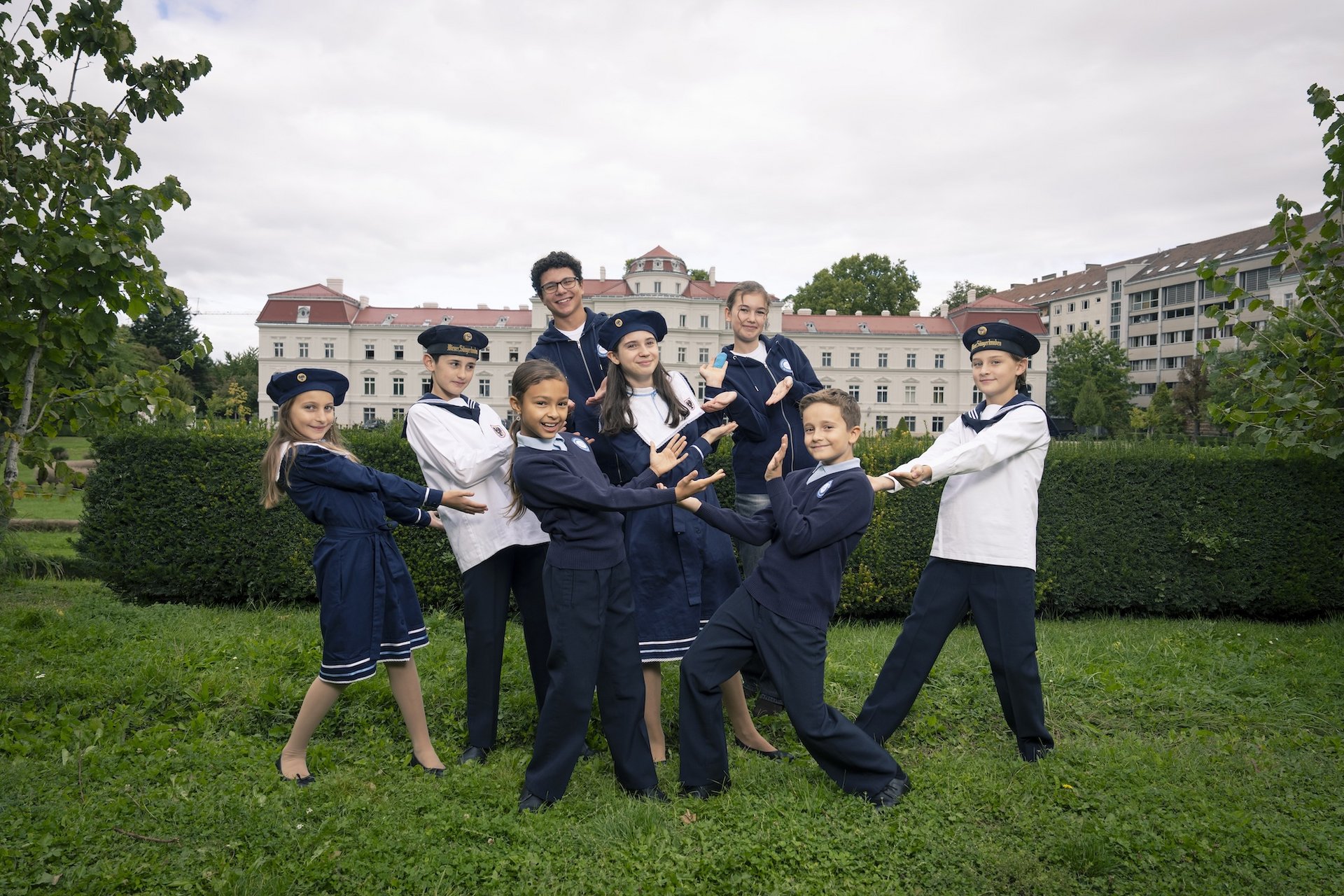 Eine Fotografie von Lukas Beck im Park des Palais Augarten. Das Bild symbolisiert die Vielfalt des Campus: Eine Gruppe von acht jungen Chormitgliedern (Mädchen und Jungen) posiert in einer spielerisch-dynamischen Formation auf einer grünen Rasenfläche. Sie tragen die verschiedenen offiziellen Outfits der Chöre – darunter dunkelblaue Matrosenkleider für die Mädchen sowie die klassischen blauen und weißen Matrosenuniformen der Jungen. Im Hintergrund erstreckt sich eine grüne Hecke, hinter der das barocke Palais Augarten unter leicht bewölktem Himmel zu sehen ist. Gemäß DSGVO werden keine Namen der Kinder genannt.