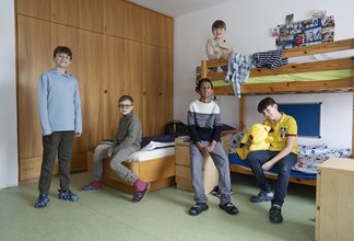 Das Foto zeigt eine Gruppe von fünf Jungen im Volksschulalter in einem hellen, freundlich gestalteten Internatszimmer auf dem Campus Augarten. Im Hintergrund ist eine große, raumhohe Schrankwand aus hellem Holz zu sehen. Auf der rechten Seite steht ein stabiles Hochbett aus Massivholz, auf dessen oberer Etage ein Junge sitzt. Die anderen vier Schüler sind im Raum verteilt: Einer steht links im Vordergrund, einer sitzt auf der Kante des unteren Bettes, und zwei weitere befinden sich in der Mitte und rechts im Raum. Die Atmosphäre wirkt ungezwungen und gemeinschaftlich. Die Aufnahme besticht durch eine natürliche Belichtung und fängt den echten Alltag im Internat ein. Urheberrecht: © Lukas Beck.