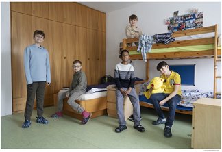 Das Foto zeigt eine Gruppe von fünf Jungen im Volksschulalter in einem hellen, freundlich gestalteten Internatszimmer auf dem Campus Augarten. Im Hintergrund ist eine große, raumhohe Schrankwand aus hellem Holz zu sehen. Auf der rechten Seite steht ein stabiles Hochbett aus Massivholz, auf dessen oberer Etage ein Junge sitzt. Die anderen vier Schüler sind im Raum verteilt: Einer steht links im Vordergrund, einer sitzt auf der Kante des unteren Bettes, und zwei weitere befinden sich in der Mitte und rechts im Raum. Die Atmosphäre wirkt ungezwungen und gemeinschaftlich. Die Aufnahme besticht durch eine natürliche Belichtung und fängt den echten Alltag im Internat ein. Urheberrecht: © Lukas Beck.