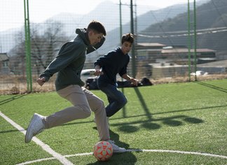 Eine Fotografie von Lukas Beck, aufgenommen im Januar 2025 während der Südkorea-Tournee der Wiener Sängerknaben. Das Bild zeigt eine sportliche Freizeitszene: Zwei Sängerknaben in dunklen Winterjacken und Hosen spielen voller Elan Fußball auf einem Rasensportplatz. Der Junge im Vordergrund ist in einer dynamischen Laufbewegung am Ball, während sein Mitschüler im Hintergrund versucht, ihn zu verteidigen. Im Hintergrund erstreckt sich eine leicht neblige Berglandschaft mit Masten. Gemäß DSGVO werden keine Namen der Kinder genannt.