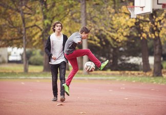 Ein Junge vollführt einen Fußballtrick im Sprung, ein zweiter Junge schaut ihm dabei zu. Die beiden befinden sich auf einem Tartan-Sportplatz, das Laub der Bäume färbt sich bereits Gelb.