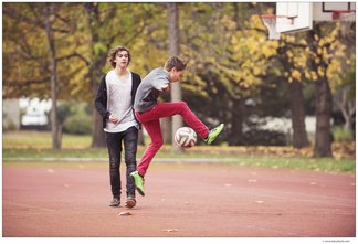 Ein Junge vollführt einen Fußballtrick im Sprung, ein zweiter Junge schaut ihm dabei zu. Die beiden befinden sich auf einem Tartan-Sportplatz, das Laub der Bäume färbt sich bereits Gelb.