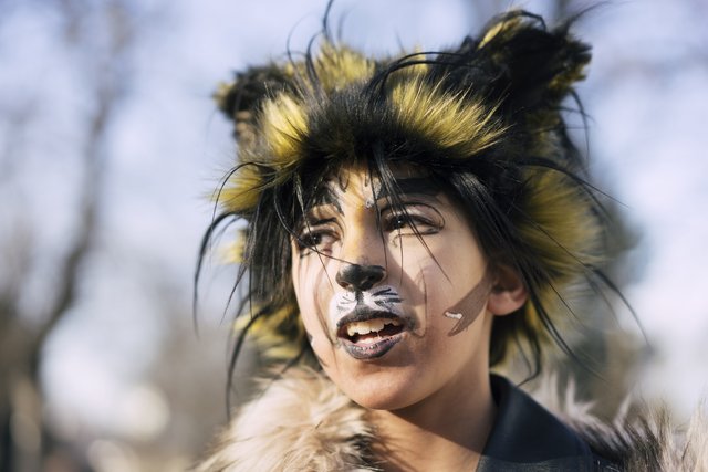 Eine Fotografie von Lukas Beck im Park des Palais Augarten während des Faschingsfests. Das Porträt zeigt ein junges Chormädchen in einer besonders aufwendigen Maskeraden-Rolle: Sie hat sich in Eigenregie als der Charakter „Rum Tum Tugger“ aus dem Musical Cats verkleidet und geschminkt. Das Mädchen blickt mit leicht geöffnetem Mund aufmerksam nach links oben. Ihr Gesicht ist detailreich geschminkt mit einer weißen Katzenschnauze, einer schwarzen Nase, feinen Schnurrhaaren und dramatischen Katzenaugen. Auf dem Kopf trägt sie die wilde schwarz-gelb melierte Fellmähne. Um den Hals liegt ein heller Pelzkragen. Aus DSGVO-Gründen wird der Name der Schülerin nicht genannt.