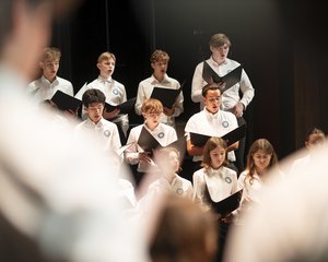 Die atmosphärische Aufnahme zeigt eine Gruppe von Jugendlichen des Chorus Juventus während einer Generalprobe im Konzertsaal MuTh. Im Bildzentrum stehen mehrere Sängerinnen und Sänger in weißen Oberteilen im hellen Scheinwerferlicht der Bühne. Sie halten geöffnete, schwarze Notenmappen in den Händen und singen hochkonzentriert. Die Gruppe ist gestaffelt aufgestellt: In den vorderen Reihen befinden sich vorwiegend die Mädchen, dahinter die Burschen des Oberstufenrealgymnasiums. Der Vordergrund des Bildes wird links und rechts von den stark unscharfen Silhouetten der Arme des Dirigenten eingerahmt, was eine dynamische Perspektive erzeugt. Der Hintergrund der Bühne ist dunkel gehalten, wodurch der Fokus ganz auf dem sängerischen Einsatz der Jugendlichen liegt. Urheberrecht: © Lukas Beck.