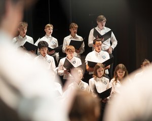 Die atmosphärische Aufnahme zeigt eine Gruppe von Jugendlichen des Chorus Juventus während einer Generalprobe im Konzertsaal MuTh. Im Bildzentrum stehen mehrere Sängerinnen und Sänger in weißen Oberteilen im hellen Scheinwerferlicht der Bühne. Sie halten geöffnete, schwarze Notenmappen in den Händen und singen hochkonzentriert. Die Gruppe ist gestaffelt aufgestellt: In den vorderen Reihen befinden sich vorwiegend die Mädchen, dahinter die Burschen des Oberstufenrealgymnasiums. Der Vordergrund des Bildes wird links und rechts von den stark unscharfen Silhouetten der Arme des Dirigenten eingerahmt, was eine dynamische Perspektive erzeugt. Der Hintergrund der Bühne ist dunkel gehalten, wodurch der Fokus ganz auf dem sängerischen Einsatz der Jugendlichen liegt. Urheberrecht: © Lukas Beck.