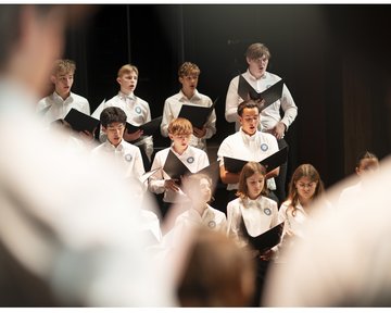 Die atmosphärische Aufnahme zeigt eine Gruppe von Jugendlichen des Chorus Juventus während einer Generalprobe im Konzertsaal MuTh. Im Bildzentrum stehen mehrere Sängerinnen und Sänger in weißen Oberteilen im hellen Scheinwerferlicht der Bühne. Sie halten geöffnete, schwarze Notenmappen in den Händen und singen hochkonzentriert. Die Gruppe ist gestaffelt aufgestellt: In den vorderen Reihen befinden sich vorwiegend die Mädchen, dahinter die Burschen des Oberstufenrealgymnasiums. Der Vordergrund des Bildes wird links und rechts von den stark unscharfen Silhouetten der Arme des Dirigenten eingerahmt, was eine dynamische Perspektive erzeugt. Der Hintergrund der Bühne ist dunkel gehalten, wodurch der Fokus ganz auf dem sängerischen Einsatz der Jugendlichen liegt. Urheberrecht: © Lukas Beck.