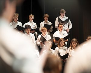 Die atmosphärische Aufnahme zeigt eine Gruppe von Jugendlichen des Chorus Juventus während einer Generalprobe im Konzertsaal MuTh. Im Bildzentrum stehen mehrere Sängerinnen und Sänger in weißen Oberteilen im hellen Scheinwerferlicht der Bühne. Sie halten geöffnete, schwarze Notenmappen in den Händen und singen hochkonzentriert. Die Gruppe ist gestaffelt aufgestellt: In den vorderen Reihen befinden sich vorwiegend die Mädchen, dahinter die Burschen des Oberstufenrealgymnasiums. Der Vordergrund des Bildes wird links und rechts von den stark unscharfen Silhouetten der Arme des Dirigenten eingerahmt, was eine dynamische Perspektive erzeugt. Der Hintergrund der Bühne ist dunkel gehalten, wodurch der Fokus ganz auf dem sängerischen Einsatz der Jugendlichen liegt. Urheberrecht: © Lukas Beck.