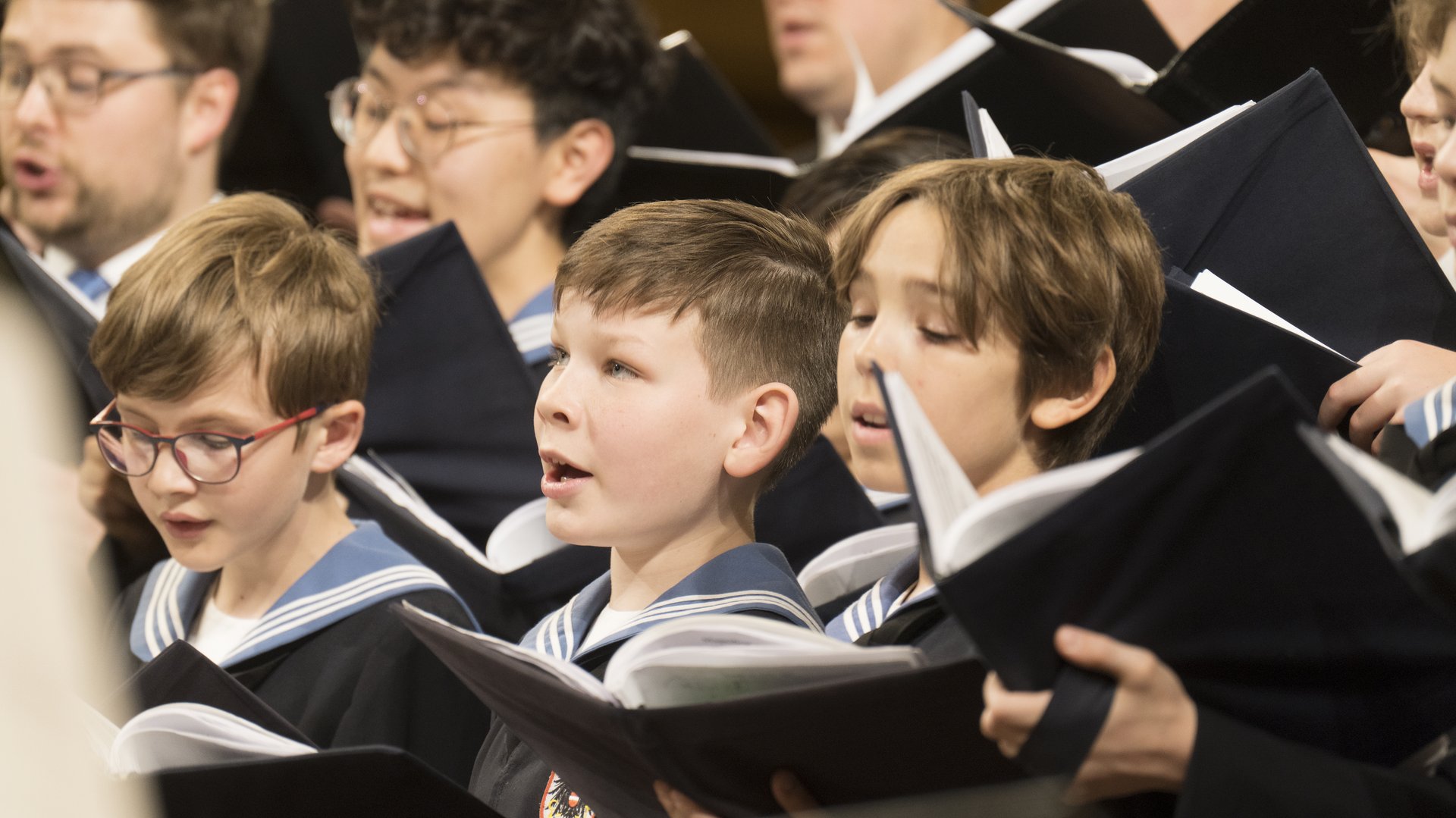 Konzertfotografie von Lukas Beck vom 525 Jahre Festkonzert der Wiener Sängerknaben. Die Aufnahme zeigt eine Gruppe junger Chorknaben in ihren traditionellen blauen Matrosenanzügen beim Singen. Im Hintergrund sind leicht verschwommen die erwachsenen Sänger des Chorus Viennensis zu sehen, die das Ensemble im Tenor und Bass unterstützen. Das Jubiläumskonzert fand unter der Leitung von Dirigent Gerald Wirth im Goldenen Saal des Wiener Musikvereins statt.

Schlagworte: Wiener Sängerknaben, Chorus Viennensis, Gerald Wirth, Lukas Beck, Festkonzert, 525 Jahre, Jubiläum, Musikverein, Goldener Saal, Knabenchor, Chormusik, Wien, Konzertfotografie