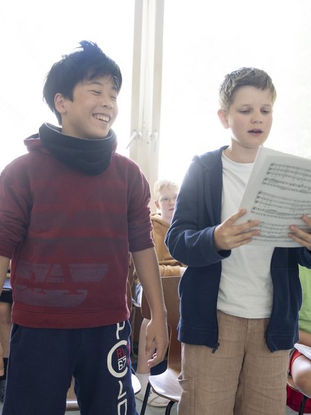 Zwei Buben stehen in einem Klassenraum singend nebeneinander. Der rechte Junge hält Noten in seinen Händen während der linke Junge lächelt.