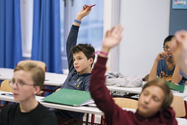 Eine Fotografie von Lukas Beck in einem Klassenzimmer der Wiener Sängerknaben. Das Bild zeigt eine lebendige Szene aus dem Unterricht: Im Vordergrund sitzen Schüler an weißen Tischen, im Zentrum streckt ein Junge im weinroten Pullover aufzeigend die Hand nach oben. Dahinter ist ein weiterer aufzeigender Schüler im grauen Oberteil zu sehen. Im linken Hintergrund hängen blaue Vorhänge am Fenster, an den Wänden sind Pinnwände erkennbar. Gemäß DSGVO werden keine Namen der Schüler genannt.