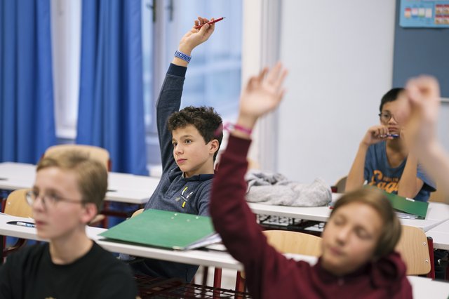 Eine Fotografie von Lukas Beck in einem Klassenzimmer der Wiener Sängerknaben. Das Bild zeigt eine lebendige Szene aus dem Unterricht: Im Vordergrund sitzen Schüler an weißen Tischen, im Zentrum streckt ein Junge im weinroten Pullover aufzeigend die Hand nach oben. Dahinter ist ein weiterer aufzeigender Schüler im grauen Oberteil zu sehen. Im linken Hintergrund hängen blaue Vorhänge am Fenster, an den Wänden sind Pinnwände erkennbar. Gemäß DSGVO werden keine Namen der Schüler genannt.