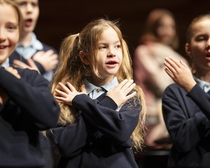Die lebendige Aufnahme zeigt eine Gruppe von Kindern des Chorus Primus während einer Aufführung im Konzertsaal MuTh. Im Vordergrund stehen drei Volksschulkinder in dunkelblauen Pullovern und hellen Hemdkragen im hellen Rampenlicht. Sie singen mit geöffneten Mündern und führen synchron eine Geste aus, bei der sie die Hände flach über der Brust gekreuzt auf die Schultern legen: die Choreografie zum Torres Straits-Song "Sessere Eye" (Eisvogel). Das Mädchen links im Vordergrund lächelt dabei fröhlich. Im Zentrum steht ein Mädchen mit langen, blonden Haaren, die zu zwei Zöpfen gebunden sind, und blickt konzentriert nach vorne. Rechts daneben singt ein weiteres Mädchen mit streng zurückgebundenen Haaren mit geschlossenen Augen. Der Hintergrund verläuft in eine warme, stimmungsvolle Unschärfe, in der weitere Chormitglieder auf den Chorrängen zu erahnen sind. Urheberrecht: © Lukas Beck.