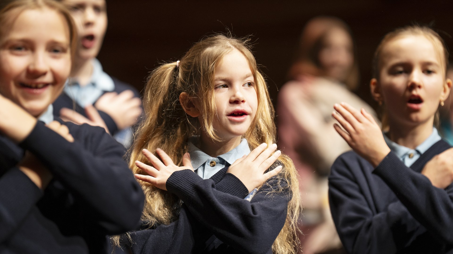 Die lebendige Aufnahme zeigt eine Gruppe von Kindern des Chorus Primus während einer Aufführung im Konzertsaal MuTh. Im Vordergrund stehen drei Volksschulkinder in dunkelblauen Pullovern und hellen Hemdkragen im hellen Rampenlicht. Sie singen mit geöffneten Mündern und führen synchron eine Geste aus, bei der sie die Hände flach über der Brust gekreuzt auf die Schultern legen: die Choreografie zum Torres Straits-Song "Sessere Eye" (Eisvogel). Das Mädchen links im Vordergrund lächelt dabei fröhlich. Im Zentrum steht ein Mädchen mit langen, blonden Haaren, die zu zwei Zöpfen gebunden sind, und blickt konzentriert nach vorne. Rechts daneben singt ein weiteres Mädchen mit streng zurückgebundenen Haaren mit geschlossenen Augen. Der Hintergrund verläuft in eine warme, stimmungsvolle Unschärfe, in der weitere Chormitglieder auf den Chorrängen zu erahnen sind. Urheberrecht: © Lukas Beck.