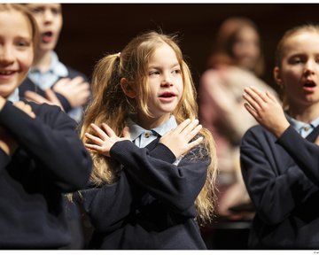 Die lebendige Aufnahme zeigt eine Gruppe von Kindern des Chorus Primus während einer Aufführung im Konzertsaal MuTh. Im Vordergrund stehen drei Volksschulkinder in dunkelblauen Pullovern und hellen Hemdkragen im hellen Rampenlicht. Sie singen mit geöffneten Mündern und führen synchron eine Geste aus, bei der sie die Hände flach über der Brust gekreuzt auf die Schultern legen: die Choreografie zum Torres Straits-Song "Sessere Eye" (Eisvogel). Das Mädchen links im Vordergrund lächelt dabei fröhlich. Im Zentrum steht ein Mädchen mit langen, blonden Haaren, die zu zwei Zöpfen gebunden sind, und blickt konzentriert nach vorne. Rechts daneben singt ein weiteres Mädchen mit streng zurückgebundenen Haaren mit geschlossenen Augen. Der Hintergrund verläuft in eine warme, stimmungsvolle Unschärfe, in der weitere Chormitglieder auf den Chorrängen zu erahnen sind. Urheberrecht: © Lukas Beck.