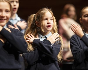 Die lebendige Aufnahme zeigt eine Gruppe von Kindern des Chorus Primus während einer Aufführung im Konzertsaal MuTh. Im Vordergrund stehen drei Volksschulkinder in dunkelblauen Pullovern und hellen Hemdkragen im hellen Rampenlicht. Sie singen mit geöffneten Mündern und führen synchron eine Geste aus, bei der sie die Hände flach über der Brust gekreuzt auf die Schultern legen: die Choreografie zum Torres Straits-Song "Sessere Eye" (Eisvogel). Das Mädchen links im Vordergrund lächelt dabei fröhlich. Im Zentrum steht ein Mädchen mit langen, blonden Haaren, die zu zwei Zöpfen gebunden sind, und blickt konzentriert nach vorne. Rechts daneben singt ein weiteres Mädchen mit streng zurückgebundenen Haaren mit geschlossenen Augen. Der Hintergrund verläuft in eine warme, stimmungsvolle Unschärfe, in der weitere Chormitglieder auf den Chorrängen zu erahnen sind. Urheberrecht: © Lukas Beck.