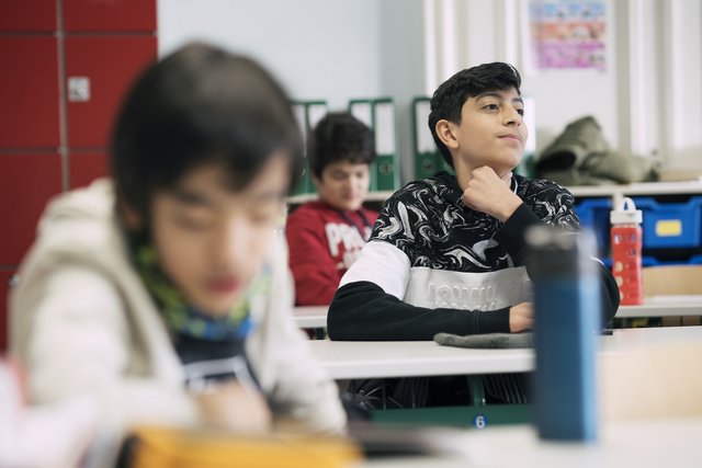 Eine Fotografie von Lukas Beck in einem Klassenzimmer der Wiener Sängerknaben. Der Bildaufbau nutzt eine selektive Schärfe: Im scharfen Mittelgrund sitzt ein dunkelhaariger Schüler in einem schwarz-weiß gemusterten Pullover und blickt aufmerksam in Richtung Tafel. Vor ihm sitzt ein Schüler im unscharfen Vordergrund, im Hintergrund ist ein weiterer Junge in einem roten Oberteil sowie Ordnerregale und ein roter Schrank zu sehen. Gemäß DSGVO werden keine Namen der Schüler genannt.