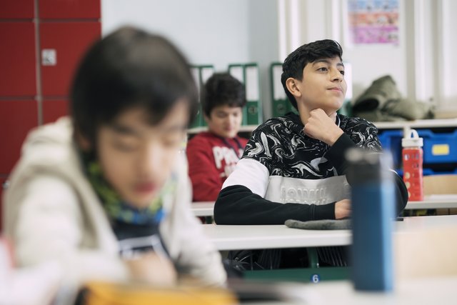 Eine Fotografie von Lukas Beck in einem Klassenzimmer der Wiener Sängerknaben. Der Bildaufbau nutzt eine selektive Schärfe: Im scharfen Mittelgrund sitzt ein dunkelhaariger Schüler in einem schwarz-weiß gemusterten Pullover und blickt aufmerksam in Richtung Tafel. Vor ihm sitzt ein Schüler im unscharfen Vordergrund, im Hintergrund ist ein weiterer Junge in einem roten Oberteil sowie Ordnerregale und ein roter Schrank zu sehen. Gemäß DSGVO werden keine Namen der Schüler genannt.